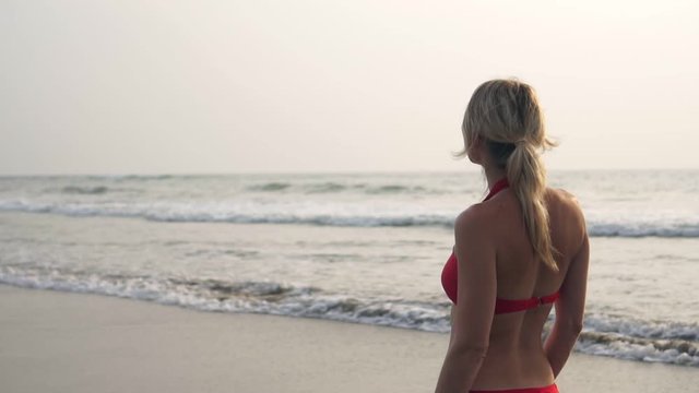 A young blonde woman in a red swimsuit walks along the sea beach, slow motion