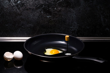 a frying egg in a heart shaped form in a frying pan on a black induction cooking. White eggshell and one egg are on the left side to the pan. A black marble-like wall as a background.
