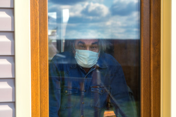 An elderly man looks out the window and wears a protective mask. The sick man is isolated to prevent infection and is quarantined at home.