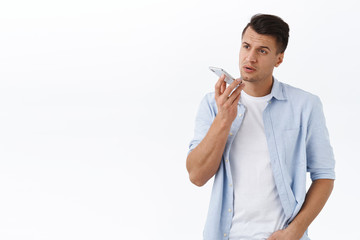 Handsome serious-looking macho man in shirt, record voice message or memo in smartphone app, holding mobile phone near lips close to dynamic, talking to person assistant, white background