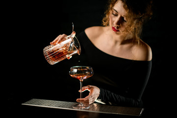 bartender girl pours finished cold cocktail from mixing cup into glass