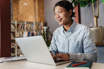 Image of joyful handsome asian man smiling and studying with laptop