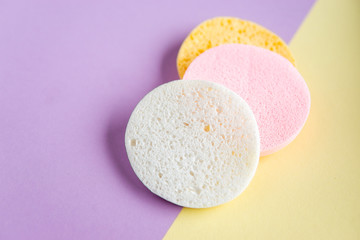 Colorful round facial  cleansing sponges on pastel yellow and purple background. Compressed natural cellulose sponges. Exfoliating Facial SPA massage. Top view, flat lay, copy space, close up.