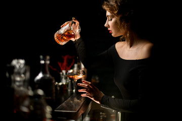 attractive woman bartender in dark bar gently pours cocktail into wineglass.
