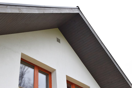 Part of white house with new eaves made of dark brown wooden planks and orange windows with ventilation grid on decorative plaser facade.
