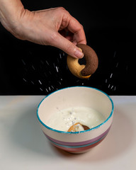 A female hand throws two-tone round biscuits into a cup of milk, splashing the liquid, on a black and white background