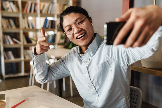Image of smiling asian man taking selfie on cellphone pointing finger