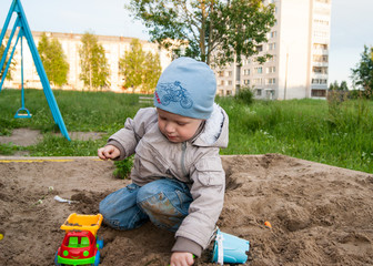 A fair-haired boy of three years in a blue hat and jeans, in a light jacket plays in the sandbox...