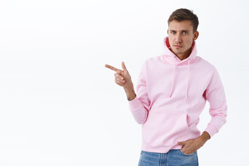 Skeptical handsome blond man look with disbelief and hesitation, pointing finger left and frowning disappointed, asking question about strange product, standing white background