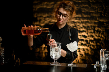 young nice girl barman pours drink from bottle into jigger.