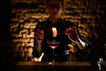 Girl bartender holds tweezers with burning plant over glass.