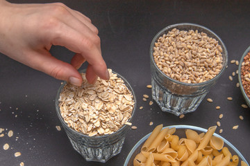 Hand reaches for dry oatmeal, which is standing on a gray background along with other cereals. Pearl barley. Buckwheat. Pasta. Vegetarian food.  Diet. Good nutrition. Healthy food.