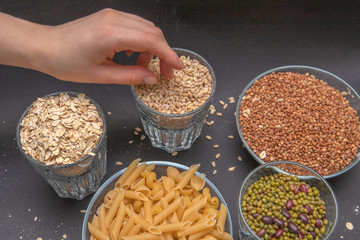 Hand reaches for dry pearl barley, which stands on a gray background along with other cereals. Rolled oats. Buckwheat. Pasta. Beans. Mash. Diet. Good nutrition. Healthy food.