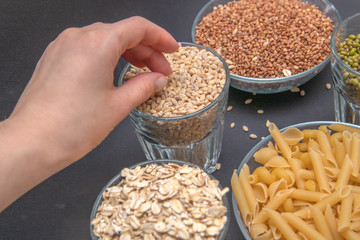 Hand reaches for  dry pearl barley, which stands on a gray background along with other cereals. Buckwheat. Pasta. Beans. Mash. Vegetarian food. Diet. Good nutrition. Healthy food. Close up.