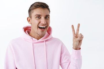 Portrait of handsome blond young man, wearing pink hoodie, staying on bright side, being positive, show peace goodwill sign and smiling broadly, enjoying amazing party, standing white background