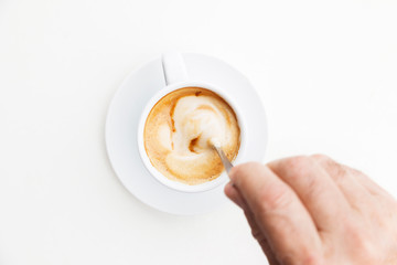 Top view of unrecognizable man dissolving sugar with spoon on cup of coffee. Isolated hot cappuccino with foam on white background. Hot drinks with caffeine concept.
