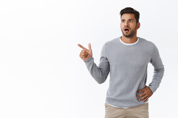 Shocked slightly worried handsome bearded caucasian guy in grey trendy sweater, gasping drop jaw from amazement, look concern, pointing left shook and speechless, check out something unusual