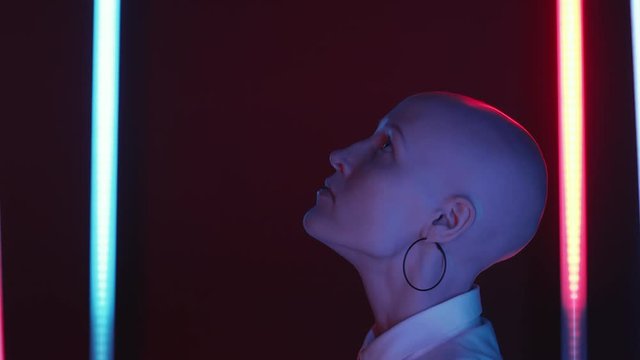 Tilt up side view shot of young bald woman in hoop earrings and white shirts standing in dark studio between two flickering neon lamps, raising her head and looking up