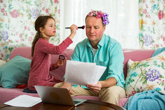 Child playing and disturbing father working remotely from home. Little girl applying makeup with brush. Man sitting on couch with laptop. Family spending time together indoors. 