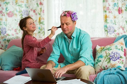 Child Playing And Disturbing Father Working Remotely From Home. Little Girl Applying Makeup With Brush. Man Sitting On Couch With Laptop. Family Spending Time Together Indoors. 