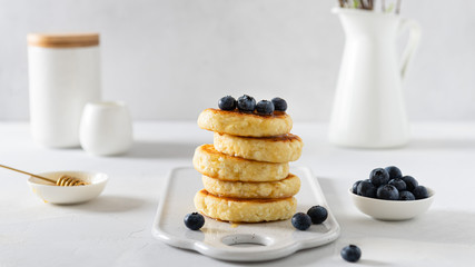 Cottage cheese pancakes, syrniki, curd fritters with fresh blueberries and honey on white background. Side view, copy space. Cookbook recipe. Russian breakfast menu