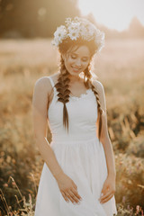 Fototapeta na wymiar portrait of smiling girl in white dress with floral wreath and braids in summer at sunset in the field