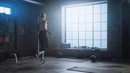 Fototapeta na wymiar Fit Athletic Blond Woman Exercises with Jumping Rope in a Loft Style Industrial Gym. She Does Her Intense Cross Fitness Training Program. Facility has Motivational Posters on the Wall.