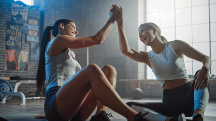 Two Beautiful Fit Athletic Girls Sit on a Floor of Industrial Loft Gym. They're Happy with their Training Program and Successfully Give a High Five.