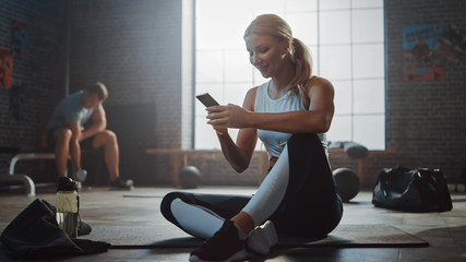 Happy and Smiling Beautiful Athletic Young Woman is Using a Smartphone while Sitting on a Floor in...