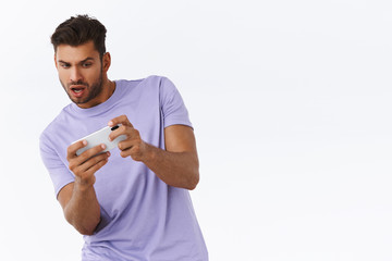 Man focused and excited with awesome new racing game, playing on smartphone, tilt body to move car on mobile phone, looking telephone thrilled and entertained, standing white background