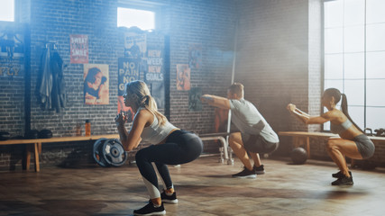 Strong Masculine Man and Two Fit Atletic Women are Doing Squat Exercises. They Workout in a Loft Gym with Motivational Posters on Walls. It's Sunny and Room has Warm Light. Part of Their Team Fitness.