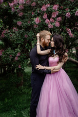 Obraz na płótnie Canvas Beautiful couple in love cuddling in a blooming lilac garden. A woman in a purple sleeveless dress with a delicate necklace around her neck. Red-haired man in a black suit kisses and hugs a girl