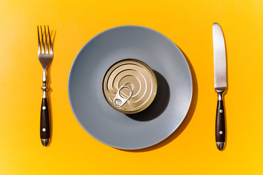 Top view of unopened can of tuna inside grey dining plate with cutlery next to it, isolated against bright yellow background.