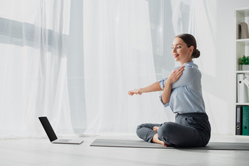Young businesswoman having online classes on laptop and stretching hands in lotus position on mat...