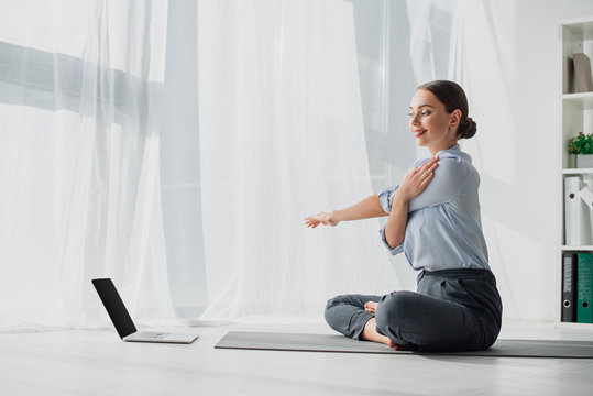 Young businesswoman having online classes on laptop and stretching hands in lotus position on mat in office