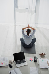 back view of businesswoman practicing yoga in lotus position with namaste gesture on mat in office...