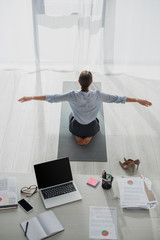 Back view of businesswoman practicing yoga in lotus position on mat in office with laptop