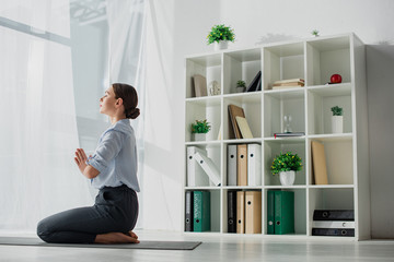 Fototapeta na wymiar Businesswoman practicing yoga and meditating with namaste gesture on mat in office