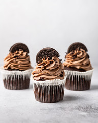 three chocolate cupcakes with cream decorated with cookies on a light background