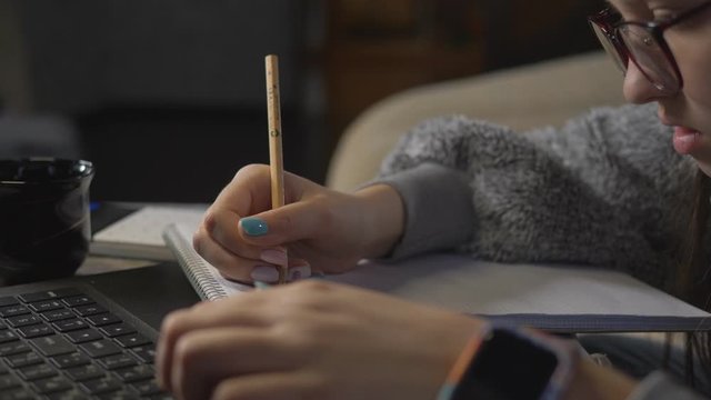Close-up, teenage girl doing homework on the distance learning front of a laptop. Girl with a pencil writing in a notebook.