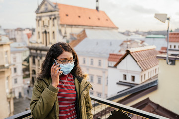 Young seriously african women wearing protective mask stands outdoors, on roof with city view on background.