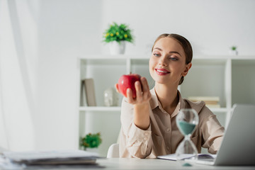 happy businesswoman holding apple at workplace with sand clock and laptop in office