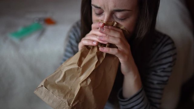 Shocked girl having panic attack staying at home in quarantine during coronavirus epidemic outbreak. Young girl breathing deeply into paper bag after watching bad news on TV about pandemic in Europe