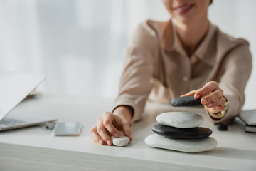 cropped view of businesswoman sitting at workplace with zen stones, smartphone and laptop