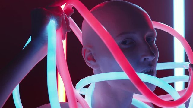 Chest-up studio shot of young beautiful woman with shaved head posing for camera in flexible neon tubes glowing with blue and pink lights