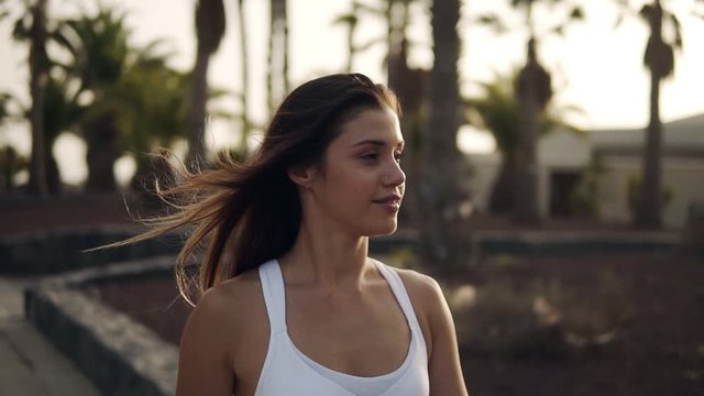 Gorgeous scene of a beautiful sporty girl walking by street beyond palm trees around. Calm walk by street, carefee girl smilling walks around. Wind blowing her hair