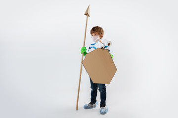 Little caucasian boy as a warrior in fight with coronavirus pandemic, with a shield, a spear and a toilet paper bandoleer. Teenboy in war for human lives. Concept of childhood, health, winning.