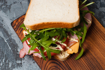 Very tasty big sandwich with ham or balyk, arugula, cheese, tomatoes and mayonnaise on a cutting board in rustic style on a gray background view from the top, copy space