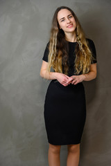 Vertical photo of a slender young Caucasian girl model with long curly hair in a black dress on a gray background. Made in the studio.