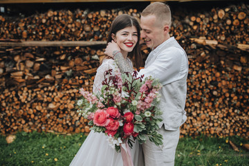Spring wedding in the mountains. A guy in a shirt and vest and a girl in a white dress and with a bouquet in their hands are standing near a barn with firewood